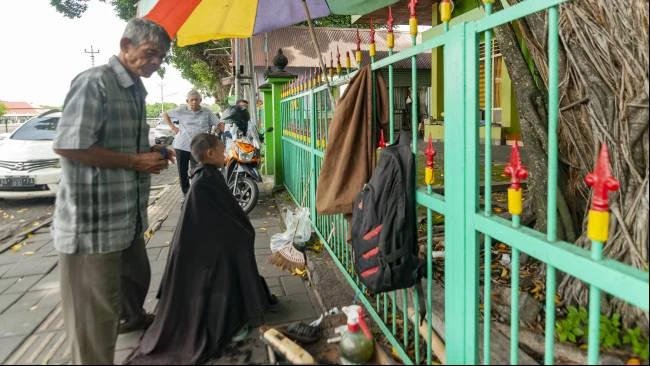Seorang pelanggan memperhatikan Udin saat mencukur cucunya, di kawasan Alun-Alun Utara, Kota Yogyakarta, Minggu (30/3/2025).