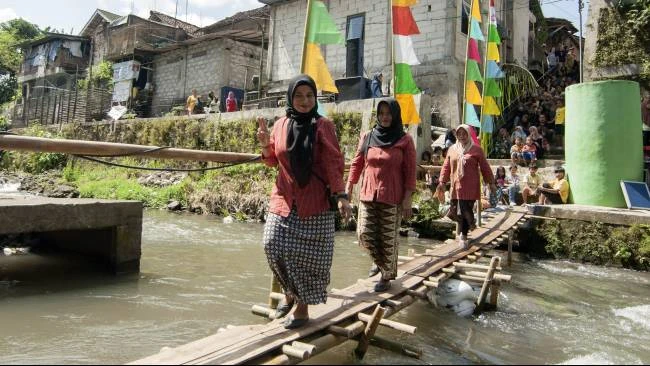 Beberapa warga melintasi jembatan bambu menuju lokasi pelaksanaan tradisi Merti Mbelik,di Tepi Sungai Winongo, Kampung Badran, Bumijo, Kecamatan Jetis, Kota Yogyakarta, Kamis (10/7/2025). 